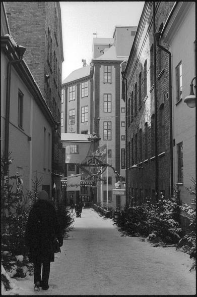 A black and white photograph of a snowy street between two large brick-buildings. There are some christmas decorations in view, for example a few christmas-trees lining the street. In the distance, a bridge over to a tall brick building in an old industrial style can be seen. There are several signs promoting the different museums housed within.