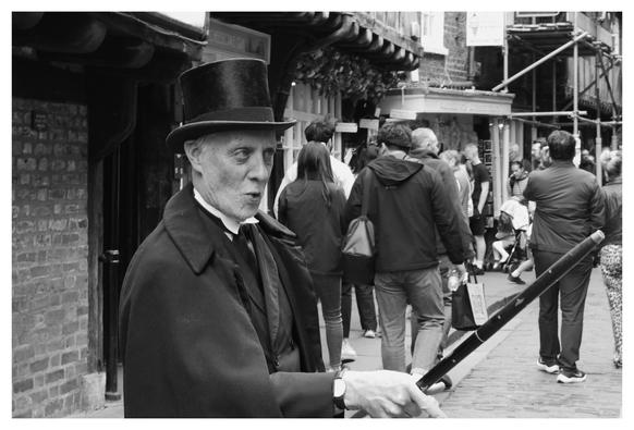 This black-and-white photograph captures a lively street scene. In the foreground, an older man dressed in Victorian-era attire stands out. He wears a tall, black top hat and a long coat, holding a cane in his right hand. His expression is serious, and he appears to be in character, possibly a street performer or historical reenactor. Behind him, a crowd of people walks along a cobblestone street lined with brick buildings. The atmosphere is bustling, with people engaged in various activities, adding a sense of movement and energy to the scene.