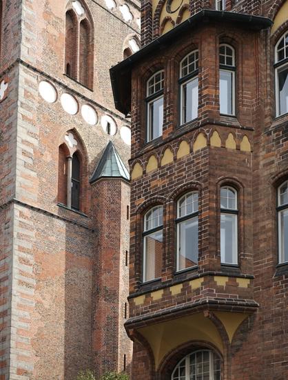 Photo montrant à gauche un pan de façade d’église gothique en brique et à droite une façade à logette également en brique mais du début du XIXe siècle.