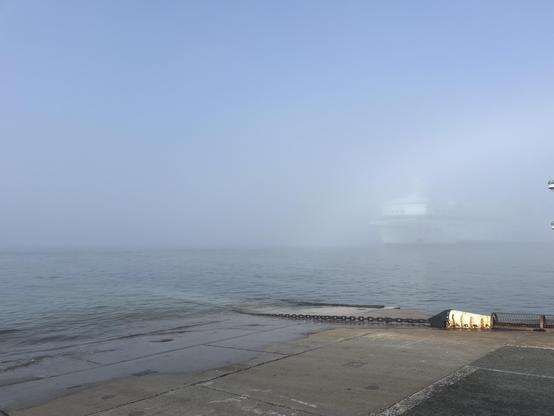 A foggy scene the chain ferry, with a large ship (Barfleur) partially obscured by mist. The calm water reflects the muted colors of the atmosphere, while the foreground features a concrete surface and a chain.