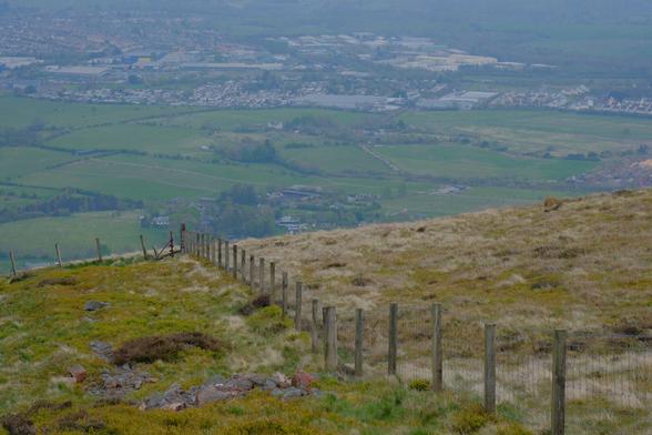 Shot from high on a hill following a fence line to the edge. Beyond and below are fields and a small town.