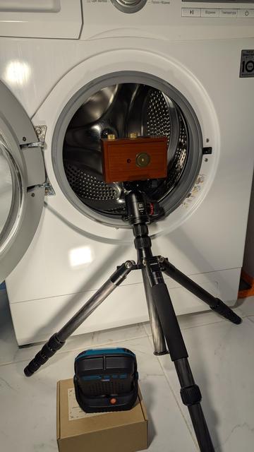 In the image, there is a wooden pinhole camera mounted on a tripod. The camera is positioned in front of an open washing machine, aiming directly into the drum. The scene appears to be set up for a creative photoshoot, capturing the inside of the washing machine. On the floor in front of the tripod, there is a small torch placed on a cardboard box. The lighting highlights the setup, creating a focused and intentional composition.