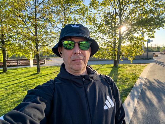 Selfie mit Hut, Sonnenbrille und Trainingjacke vor einer Wiese mit Bäumen  und der Sonne zwischen den Bäumen.