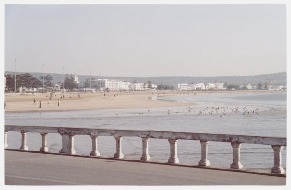 Photo of a serene beach scene featuring a sandy shoreline, a calm ocean with birds, and a damaged but still charming stone balustrade in the foreground. People are walking along the beach and near the water, with white buildings visible in the background under a clear sky.