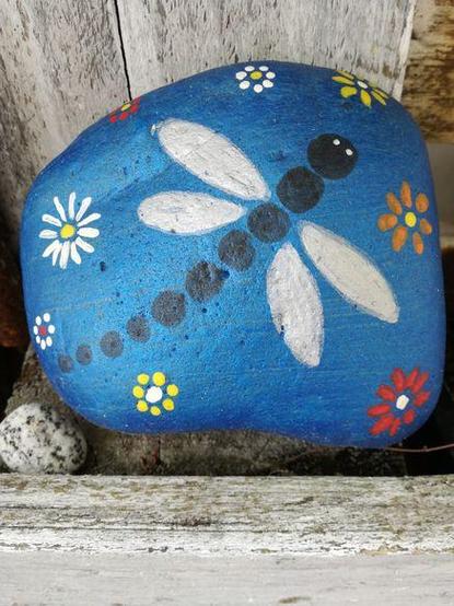 Eine Libelle mit schwarzem Leib und weißen Flügeln auf einem blau grundierten Stein .
Um die Libelle herum einige einfach gemalte Blüten in weiß rot und gelb.