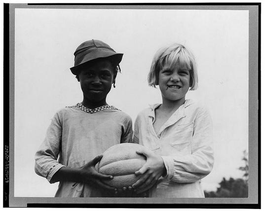 The black and white photograph depicts two children standing side by side against a plain background. The child on the left appears to be wearing a wide-brimmed hat with decorative elements around its base, possibly made of fabric or straw. They are dressed in what looks like a loose-fitting, long-sleeved top with visible buttons down the front and rolled-up sleeves.

The other child is smiling broadly at the camera, showing their teeth. This individual has short hair that appears tousled, likely blond, and they're wearing a light-colored shirt or jacket with several pockets on one side. They are holding an object in both hands, which seems to be a round ball made of leather or another material.

Both children have marks around their mouths; the left child's seem like faint lines possibly from biting down too hard while smiling, and those on the right might indicate some kind of playful activity involving food, as they could resemble smudges of chocolate or peanut butter.