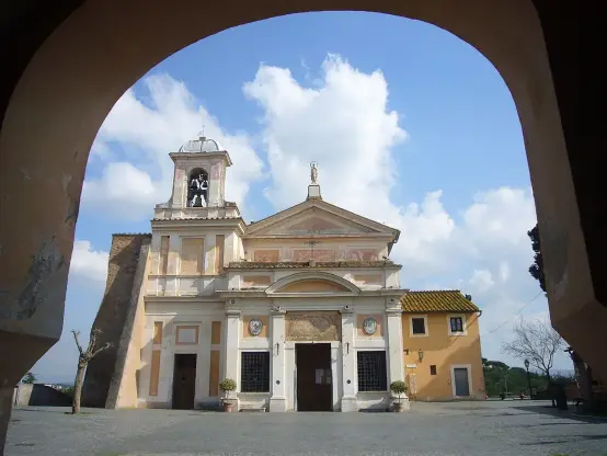The image features a church or religious building viewed through an archway. The structure has a bell tower and various architectural details, including decorative elements on the façade. The sky is bright with clouds, and there are trees and a yellow building in the background. It appears to be a picturesque scene, likely in an outdoor setting.

Image Credits: Wikimedia / Lalupa / CC BY-SA 3.0