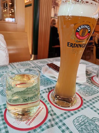 Picture of a glass mug of white Austrian wine next to a tall glass of German weissbier, set on a traditional style woven table cloth. The tablecloth is decorated with pictures of hops, wheat, beer and pretzels.