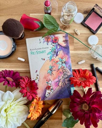Livre posé au milieu de divers éléments: fleurs de couleurs différentes, pilules et boites de pilules, ustensiles et produits de maquillage. La couverture de Re:Start montre une femme au visage coupé par l'angle choisi, tenant des fleurs en main, entourée de fleurs et de pilules médicamenteuses.
