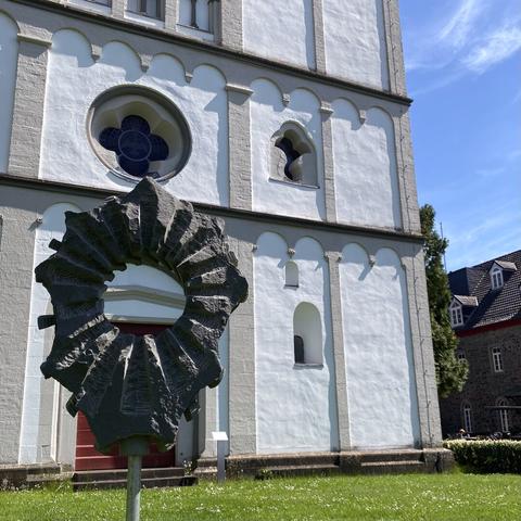 Im Vordergrund eine an eine Monstranz erinnernde Steinskulptur. Im Hintergrund die schlichte weiße Fassade einer Kirche. Ein bisschen Wiese und blauer Himmel sind auch zu sehen.