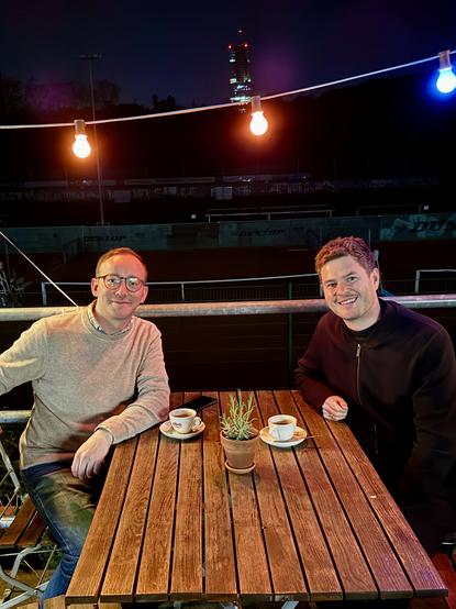 Dax und Moritz mit zwei Tassenkaffee an einem Holztisch im Freien, sie sehen glücklich aus und lächeln in die Kamera

Über ihnen bunte Glühbirnen, die ein gemütliches Licht ins Dunkel der Nacht bringen