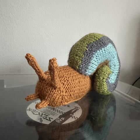 A knit snail. The body is orange. The shell is green, grey, blue, and tan.