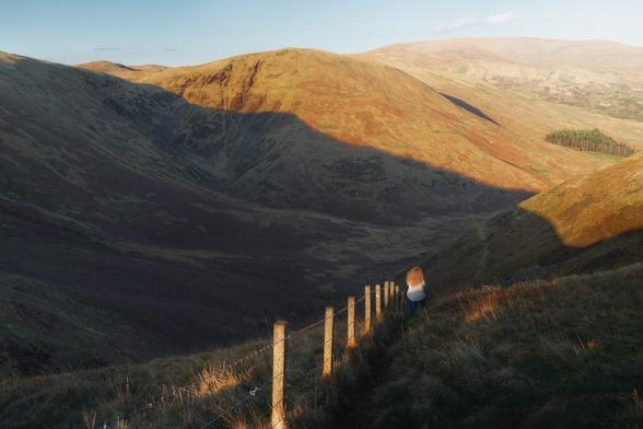 A sweeping, sunlit landscape of the Devil's Beef Tub near Moffat, Scotland, with dramatic rolling hills casting long, deep shadows across the rugged valley; in the foreground, a solitary figure with long, curly blonde hair, wearing a white top, stands next to a weathered fence, gazing thoughtfully into the vast, serene expanse under a clear blue sky.