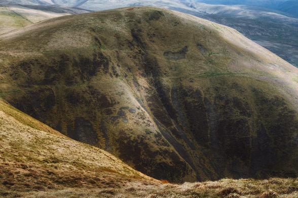 A dramatic view from Annanhead Hill above the Devil’s Beef Tub, looking towards the steep, rugged western face of Great Hill, its grassy slopes streaked with dark, weathered patches and illuminated by a soft, angled light, while distant hills, bathed in cooler tones, gently fade into the misty background.