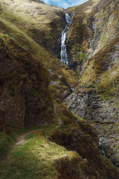 The dramatic scene of Grey Mare’s Tail near Moffat, where a tall, narrow waterfall cascades down a steep, rocky gorge surrounded by rugged, grass-covered hills, with a winding path snaking through the foreground and climbing towards the towering cliffs under a bright but partly clouded sky.