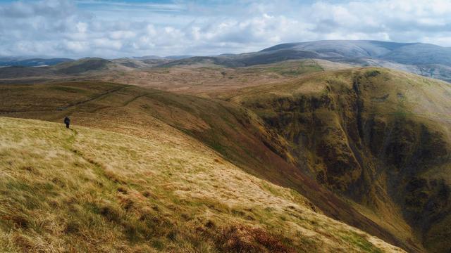 A lone walker with a backpack following a faint grassy path along the windswept slopes of Annanhead Hill, looking towards the dramatic, rugged western face of Great Hill along the Annandale Way near Moffat, Scotland, with a vast panorama of rolling hills stretching into the distance beneath a bright, cloud-streaked sky.