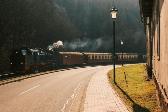 Ein Vintage-Dampfzug ist zu sehen, der entlang einer geschwungenen Straße fährt, mit Rauch, der aus der Lokomotive aufsteigt. Die Szene spielt in einem bewaldeten Gebiet, begleitet von Laternenpfosten, die den Weg säumen. Eine grasbewachsene Fläche und ein verwittertes Gebäude sind ebenfalls sichtbar