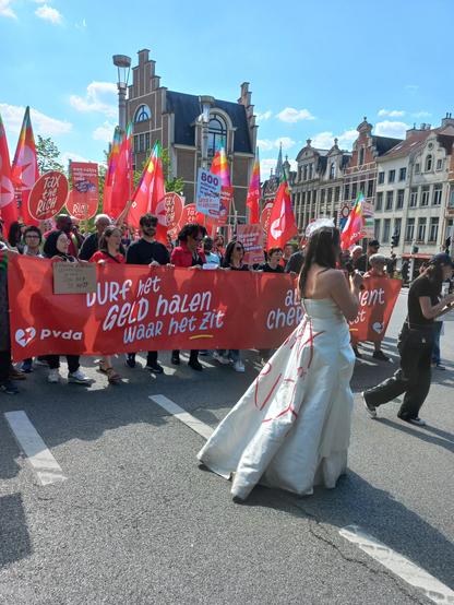 Vrouw met lange witte jurk met daarop 'Tax the rich!' die scandeert met daarachter een optocht vol rode vlaggen en een banner met daarop 'Durf het geld halen waar het zit'