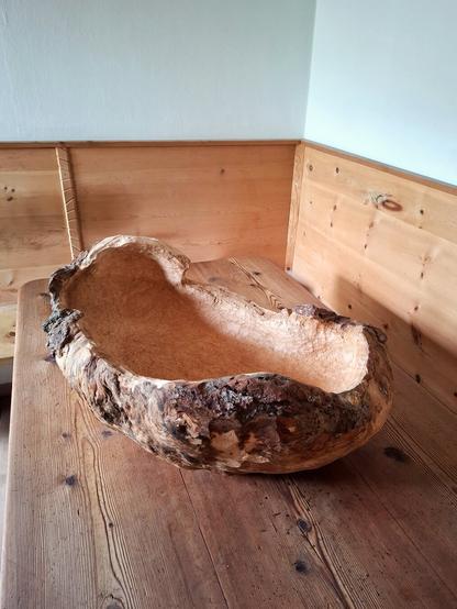 A large, rustic wooden bowl carved from a spruce burl, resting on an aged wooden larch table. The bowl has an irregular, organic shape, with a rough, textured exterior showing bark remnants and natural knots, while the interior is smoothly hollowed out. The setting is cozy with a wooden paneled walls.