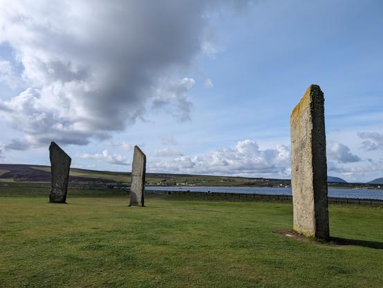 Three standing stones: one in the foreground on the right and two on the left, closer together and further from the camera. There is a body of water in the background on the right and the sky is light blue with a gray cloud on the left.