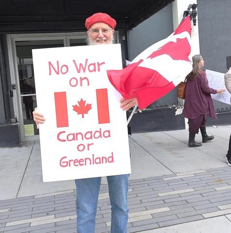 Person with Canadian flag and picket sign, "No war on Canada or Greenland".