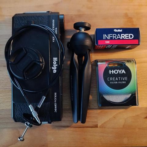 Several Items knolled on a wooden table:
* Holga 120WPC pinhole camera with lens cap
* Coiled long cable release
* Manfrotto table top folding tripod
* A box Rollei IR film
* Hoya Infrared R72 filter, in plastic case
