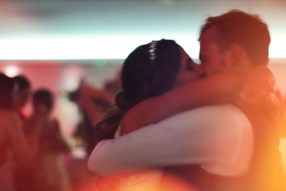 A blurred photo of a bride and groom kissing on the dance floor. The bride has a jeweled headband and her arms around the groom’s neck. The background shows wedding guests dancing, out of focus. Warm orange light flares across the image, adding a soft glow.