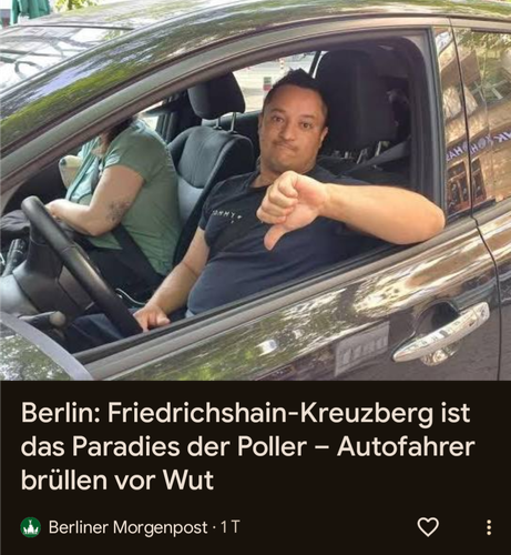 Das Bild zeigt zwei Personen in einem Auto. Der Fahrer, der im Vordergrund sitzt, trägt ein schwarzes T-Shirt mit dem Aufdruck "Tommy" und zeigt mit der rechten Hand einen Daumen nach unten. Er hat kurze, dunkle Haare und einen leicht lächelnden Gesichtsausdruck. Die Person im Hintergrund, die den Fahrersitz belegt, trägt ein grünes T-Shirt und hat ein Tattoo auf dem linken Arm. Sie blickt nach unten und scheint auf etwas zu konzentrieren. Das Auto ist ein dunkelgrauer Sedan mit einem Chromgriff am Türgriff. Im Hintergrund sind Bäume und ein Teil eines Gebäudes zu sehen. Der Text auf dem Bild lautet: "Berlin: Friedrichshain-Kreuzberg ist das Paradies der Poller – Autofahrer brüllen vor Wut" und ist von der Berliner Morgenpost.