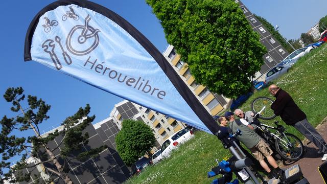 Drapeau "Réparez votre vélo avec Hérouelibre" entre bleu azur, verts vifs et anthracite des immeubles à Hérouville Saint-Clair