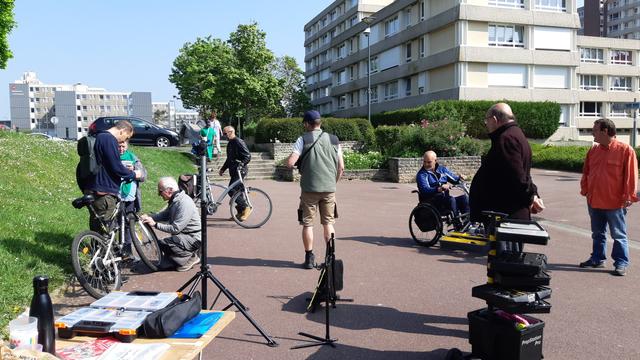 Hommes et femmes en train d'arriver, réparer et regarder l'atelier d'aide à la réparation vélos, sur une place piétonne sous un grand soleil. Boissons, gobelets, comm et outils sur une table au premier plan.