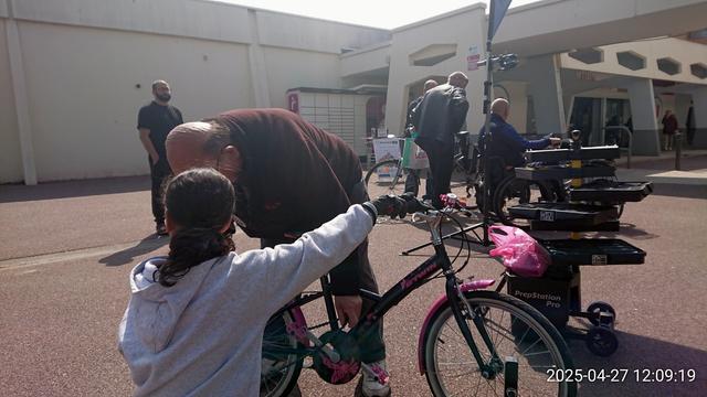 Fillette gantée et homme remettant la chaîne d'un vélo enfant en place, devant une servante mobile à outils, 4 hommes en train de converser autour du drapeau de l'association, et le panneau "Parlons-vélo.fr"