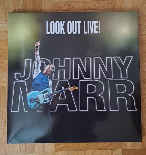 Record cover. Plain black background, except for two strobe lights, both at the top of the cover, one to the left, the other to the right. Between the lights is the text "Look out live!" and below that the text "Johnny Marr".  Standing in front of his name, but more to the right than the middle is the man himself, Johnny Marr, holding a light blue Fender guitar, with one arm reaching up to the sky.