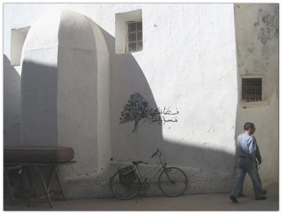 Photo of a turquoise vintage bicycle with red bag in the centre of the picture, in the shadow, standing close to a white house wall half in the sun. Over the bike on the wall is a "graffiti" or drawing of a green tree with an Arabic sentence written in black, meaning "Your cleanliness is your own responsibility". Two small windows are higher on that same wall and a third one on the right side of the picture, just around the corner of the house, over two men walking on the paved street. On the left side of the picture along the same wall is a mattress with red squares fabric on a wooden table.
