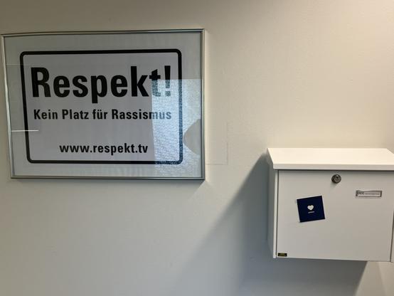 A framed poster reads "Respekt! Kein Platz für Rassismus" (Respect! No Place for Racism) alongside a white mailbox featuring a small blue card with a heart symbol. The setting appears to be an indoor space.