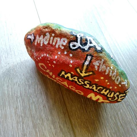 An elongated painted rock on a faux grained floor. The rock facing the viewer is painted in reds and the opposing face in greens. The front has names of places and US states, painted white, some with black outline, with some arrows and dotted meandering paths: Maine, LA, Massachusetts, Chicago, Florida, NY, RI, California.