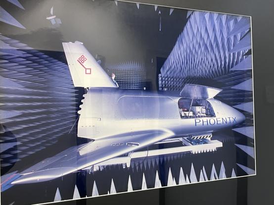 An aircraft model named "PHOENIX" is displayed in a specialized testing facility, surrounded by sound-absorbing triangular panels designed to minimize interference. A person is visible near the model, suggesting a testing or evaluation setting.