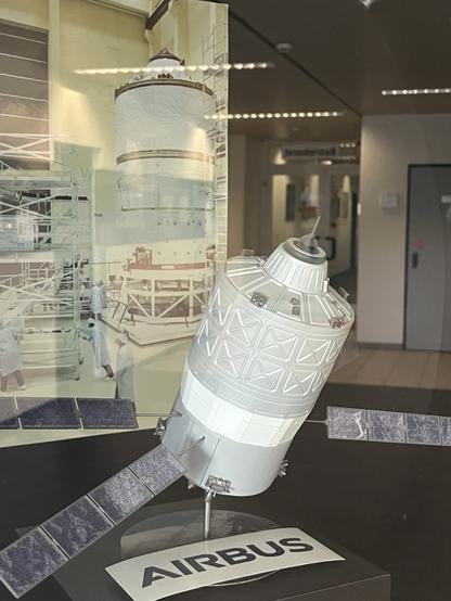 A model of a spacecraft is displayed in front of an image showcasing a spacecraft assembly area. The model features solar panels and is labeled "AIRBUS." The background image includes technicians in white clothing working on a larger assembly.
