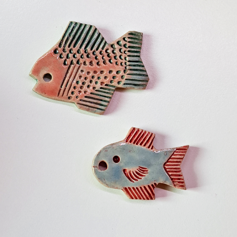 Two small ceramic fishes, one larger which is pink with bluish green fins, and the other which is blue with pink fins.