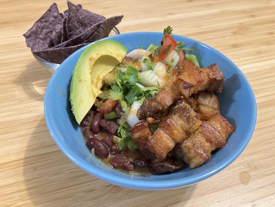 A bowl of beans, pork, salsa, pico de gallo, & avocado, with a small bowl of corn chips on the side.