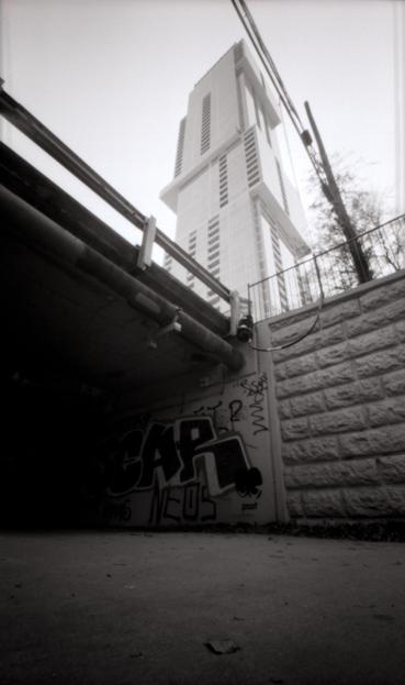 Black and white pinhole photograph of the "jenga tower" in Austin, TX. The shot is taken from below street level near a bridge. Graffiti is visible below the bridge where the sidewalk disappears into darkness.