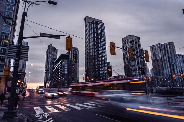 Long exposure photo of Toronto rush hour, with blurred cars and a streetcar moving through an intersection, surrounded by tall residential buildings under a gray evening sky.