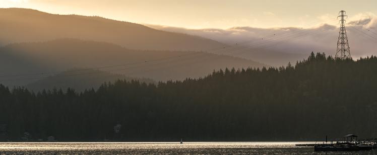 A sunrise photograph of the west side of Diez Vistas, as seen from Deep Cove. Sun is just peeking out from over the mountains in the upper left of the frame, spilling light over the water and trees of the inlet. A signal tower (which I've shot many, many times before) is visible top right, highlighted by the remaining pre-dawn clouds behind. Bottom right shows the leading edge of a nearby pier.