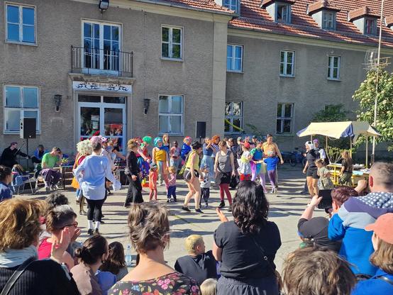 Vor einem älteren großen Haus ist eine bunte Tanzgruppe zusammen mit Kindern zu sehen, die fröhlich tanzen und Publikum, welches der Darbietung zuschaut.
