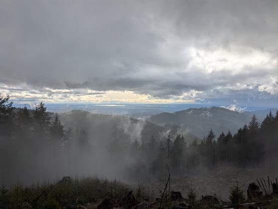 View from a mountain, with a cloudy sky. Lakes in the far background, trees in the middle ground.