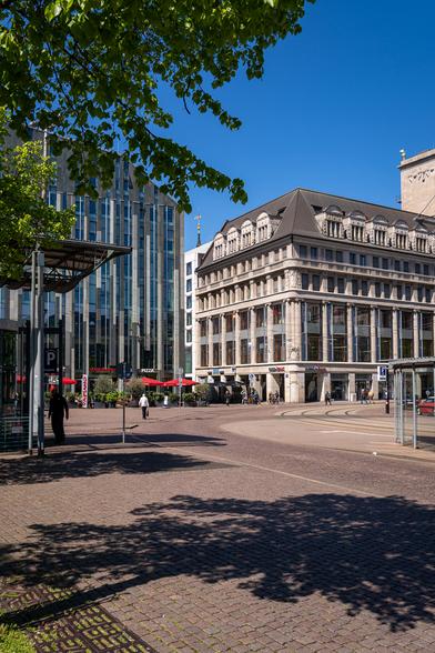 Blick über einen Platz auf Hausfassaden. Eine Baumkrone ragt ins Bild.