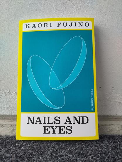 Kaori Fujinon Nails and Eyes -kirjan kansikuvassa on keltaista reunoilla ja turkoosia keskellä. Turkoosissa osassa näkyy kaksi linssinmuotoista esinettä.