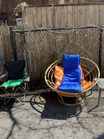 Two chairs in the sun in front of a wooden and metal fence. One black folding camp chair with a bright green seat cushion, the other a wicker round basket frame with an orange and blue make shift cushions on it