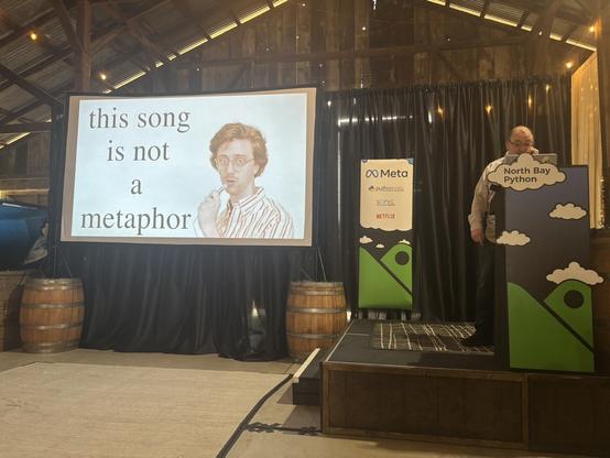 Glyph presenting on the North Bay Python stage. The slide has a picture of Brian David Gilbert singing his caterpillar song with the title "this song is not a metaphor", as Glyph realizes this is a Python conference and not a hydraulic excavator conference.
