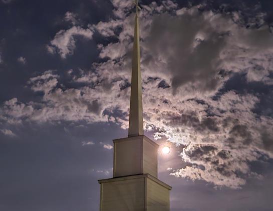Looking up into spooky night sky where the moon's glow illuminates cloud formations surrounding a tall church steeple.