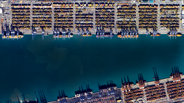 a satellite picture of a big commercial harbor filled with shipping containers and cargo ships.
there are lots of automated cranes all around the blue shipping channel taking up all the middle of the frame
